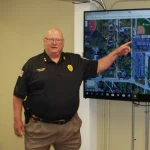 Bald man in police uniform, identified as Charles P. Garber Jr, points at a map on a large screen mounted on the wall.