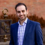 Ata Karbasi, Ph.D., is seen smiling outdoors in a blue blazer and plaid shirt, standing near a brick wall and a metal fraternity symbol.