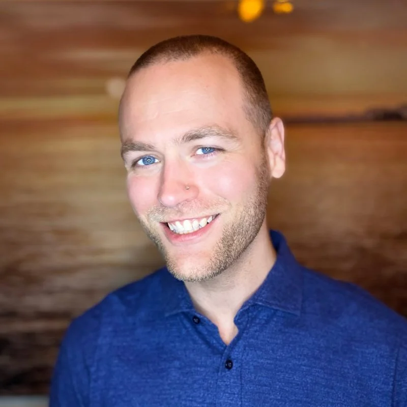 Smiling man with light eyes in a blue shirt, Bradford Thompson stands in front of a blurred, warm-toned background.