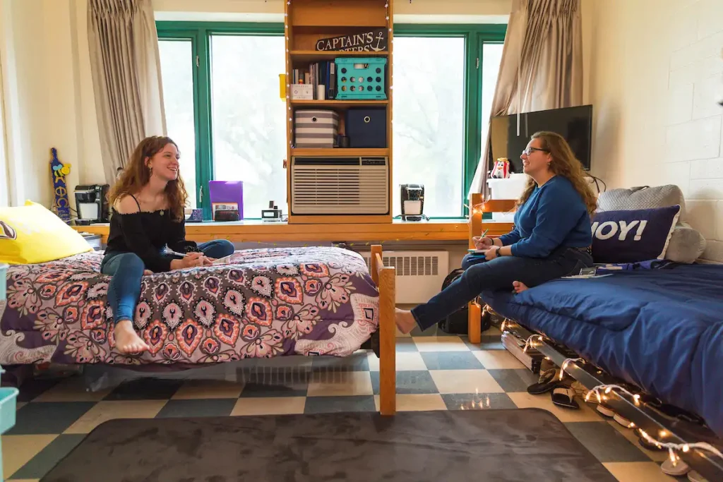 Two college students talk while sitting on separate beds in a bright, neatly organized dorm room, comparing the comforts of their current space to the options available in off-campus housings nearby.