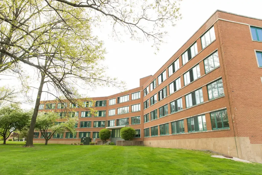 Four-story brick building with many windows, surrounded by green grass and trees under a cloudy sky, ideal for Off-Campus Housings.