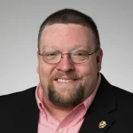 Smiling man with glasses, a beard, and short hair, wearing a suit jacket and pink shirt—Brett Calland.