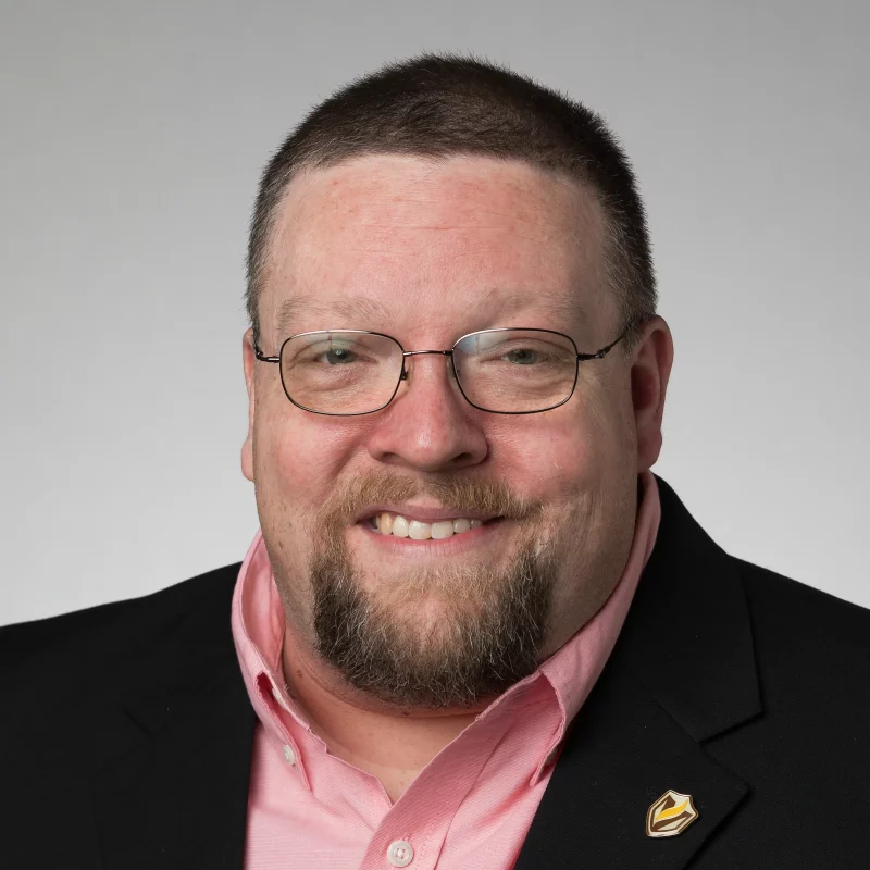 Smiling man with glasses, a beard, and short hair, wearing a suit jacket and pink shirt—Brett Calland.