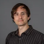 A man with brown hair wearing a black striped shirt poses in front of a plain gray background.