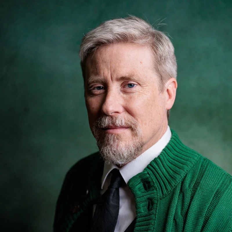 Martin Buinicki, a middle-aged man with light hair and a beard, wears a green sweater and black tie as he poses against a green background.