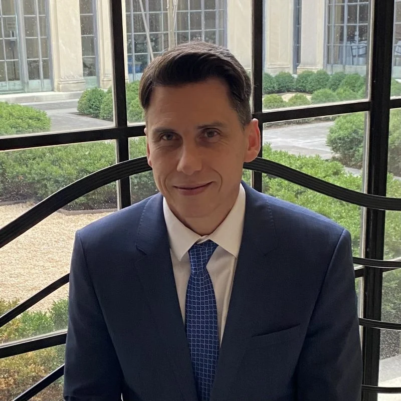 Carter Hanson, dressed in a blue suit and tie, sits indoors by large windows with a garden view in the background.