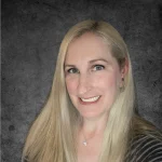 Carrie Miller, with long blonde hair, smiles warmly in a striped top and necklace against a dark textured background.