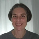 Smiling woman with short brown hair, MBA, and CPA, wearing a gray top, standing in front of white curtains.