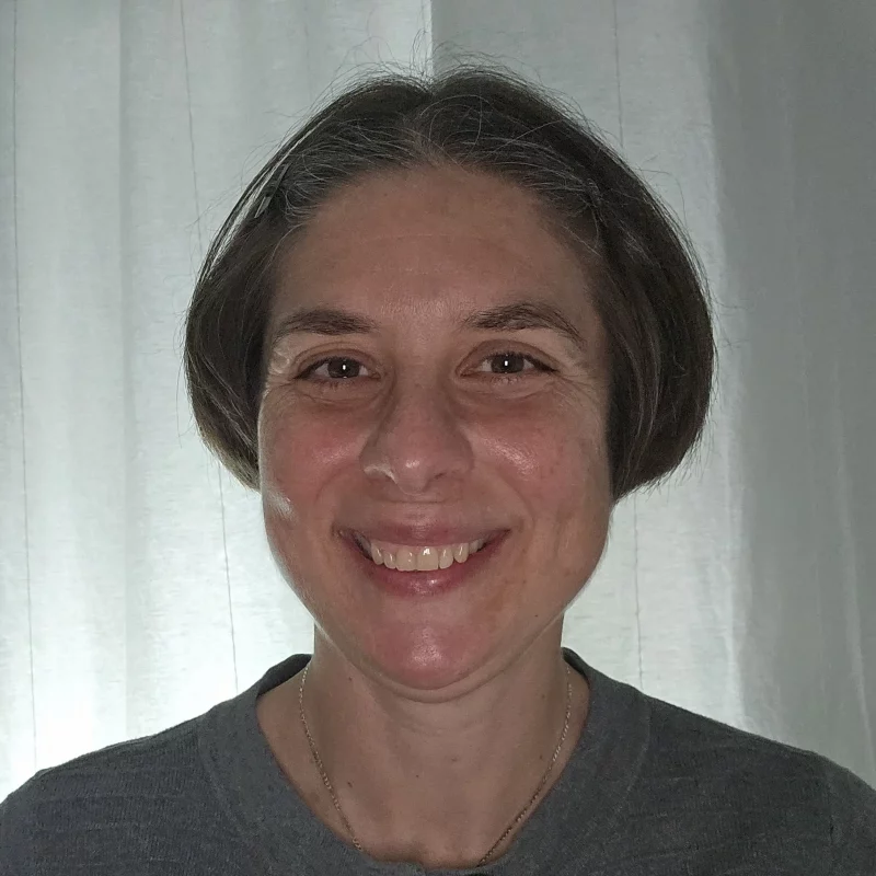 Smiling woman with short brown hair, MBA, and CPA, wearing a gray top, standing in front of white curtains.