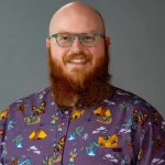 Smiling person with glasses and a long red beard, Chris Dabbs, NCC, LMHC, wearing a colorful, patterned shirt with dragons and monsters.