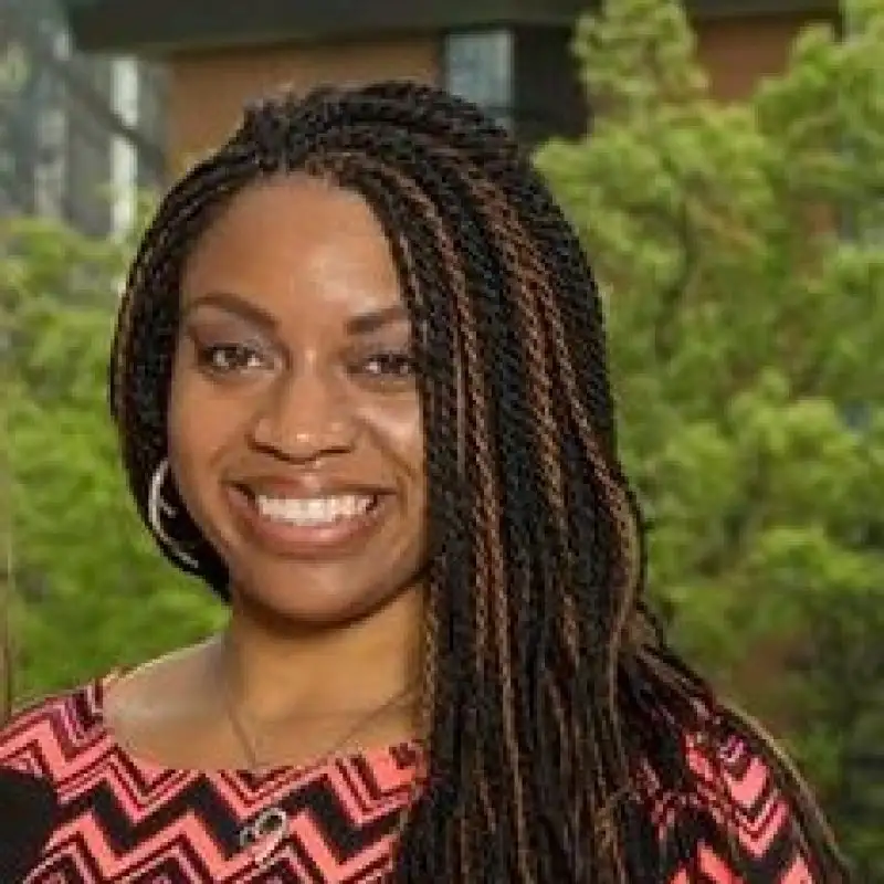 A woman with long braided hair, part of the team, smiles outdoors while wearing a pink and black patterned top.