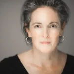 Christine Maul Rice, a woman with gray hair, wearing hoop earrings and a black top, looks into the camera with a calm expression.