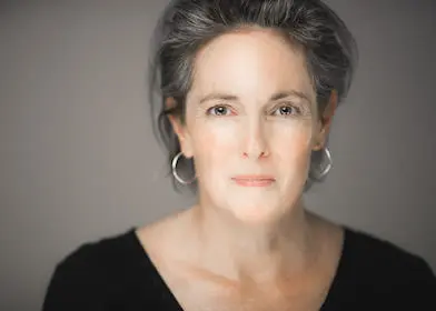 Christine Maul Rice, a woman with gray hair, wearing hoop earrings and a black top, looks into the camera with a calm expression.