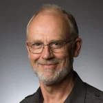 Older man with glasses, short gray hair, and beard, Chuck Schaefer is smiling at the camera against a gray background.