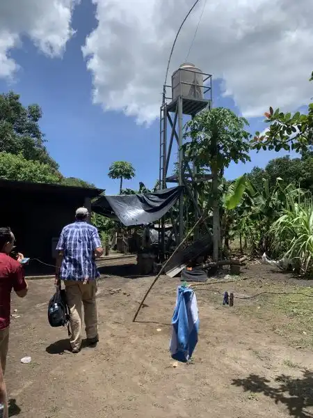Two people walk toward a tall water tank structure in a rural, tropical setting under a partly cloudy sky, as gentle WAVES of heat shimmer in the distance.