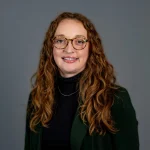 Bailey Regier, RN, MSN, with long curly hair, glasses, a black top, and dark blazer, smiles confidently in front of a gray background.