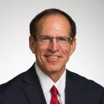 Smiling man with glasses, identified as Stewart Cooper, Ph.D., in a dark suit, white shirt, and red tie, against a plain light background.