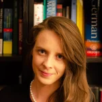 Woman with long brown hair and pearl necklace, smiling in front of bookshelves filled with books—an Assistant Professor of World Languages passionate about connecting diverse cultures.
