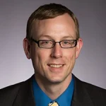 Dan White, Ph.D., is a man with short light brown hair, wearing glasses, a suit, blue shirt, and yellow patterned tie, smiling.
