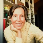 Danielle Lavin-Loucks, in a cream sweater, smiles warmly while resting her chin on her hand, seated in front of bookshelves.