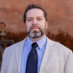 Ph.D. David Rolling, a man with short hair and beard, stands outdoors in a light suit jacket, striped shirt, and black tie.