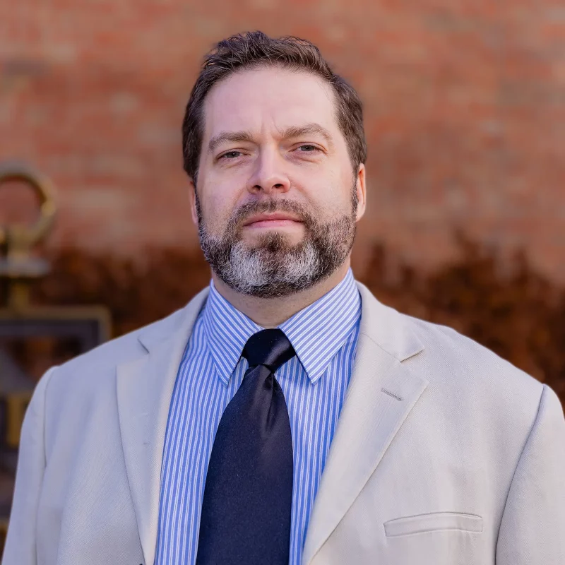 Ph.D. David Rolling, a man with short hair and beard, stands outdoors in a light suit jacket, striped shirt, and black tie.