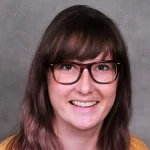 Christina M. Davis, a woman with brown hair, glasses, and a nose piercing, smiles warmly in front of a gray background.
