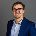 Devin Sodums, MS, is a man in glasses and a blue suit jacket smiling in front of a plain gray background.