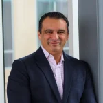 Dushan Nikolovski, dressed in a dark suit and light shirt, smiles while standing indoors by a window and a gray wall.