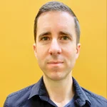 Man with short brown hair and a navy shirt standing in front of a yellow background.