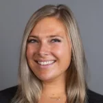 Smiling woman with straight blonde hair, wearing a black top, poses confidently against a plain gray background—Emily De Vita.