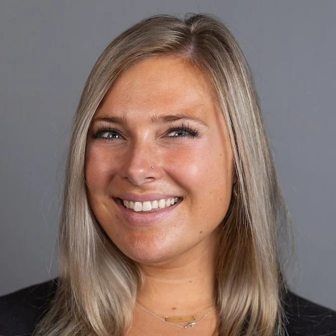 Smiling woman with straight blonde hair, wearing a black top, poses confidently against a plain gray background—Emily De Vita.