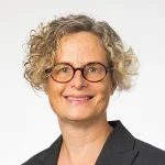 Smiling Jennifer Prough with short curly hair, glasses, and a dark blazer on a plain light background.
