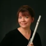 Cynthia Fudala, a woman with brown hair, holds a flute and smiles warmly in front of a dark background.