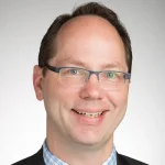 Smiling man with glasses, short brown hair, and a checkered shirt under a dark jacket, against a light background—Gregg B. Johnson, also known as Bagel Johnson.