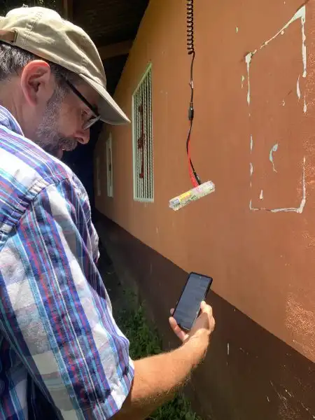 Man in a cap holding a phone, standing near a wall with a small device hanging from a wire, possibly measuring WAVES.