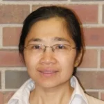 A woman with glasses and dark hair smiles softly in front of a brick wall.