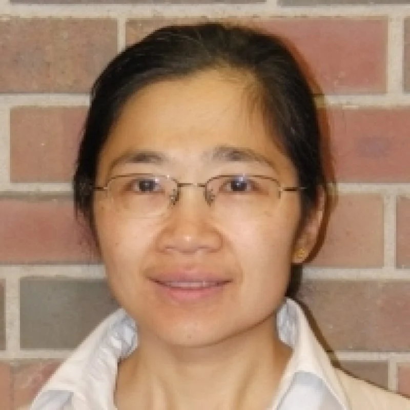 A woman with glasses and dark hair smiles softly in front of a brick wall.