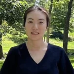 Hana Jun, a woman with dark hair in a black shirt, stands outdoors with lush green trees and grass in the background.