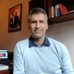 Haydar Çukurtepe, wearing a light blue sweater, sits indoors with a desk and bookshelves in the background.