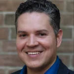 Diego Piedra, a man with short dark curly hair and a blue shirt, smiles in front of a brick wall.
