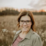 Jennifer Easthope, a woman with brown hair and glasses, smiles in a field at sunset, wearing a light jacket and pink top.
