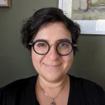 Smiling Margarita Rayzberg with short dark hair and round glasses, wearing a black top and necklace, indoors.