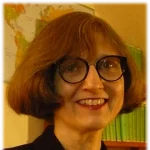Smiling woman with glasses and brown hair, Susan Holman stands in front of a map and a shelf of green books.