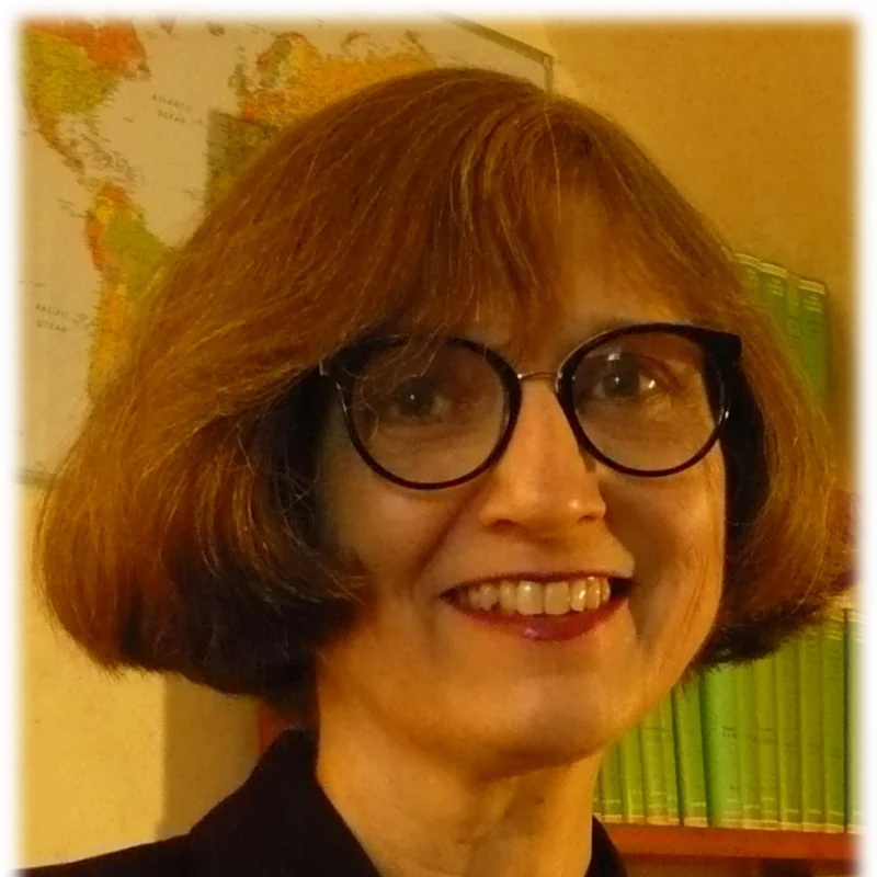 Smiling woman with glasses and brown hair, Susan Holman stands in front of a map and a shelf of green books.