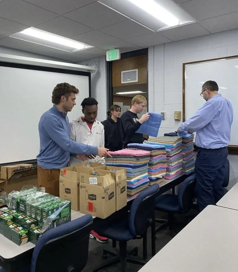 Four people stack folded towels and organize supplies in a classroom with tables, boxes, and WAVES of teamwork.