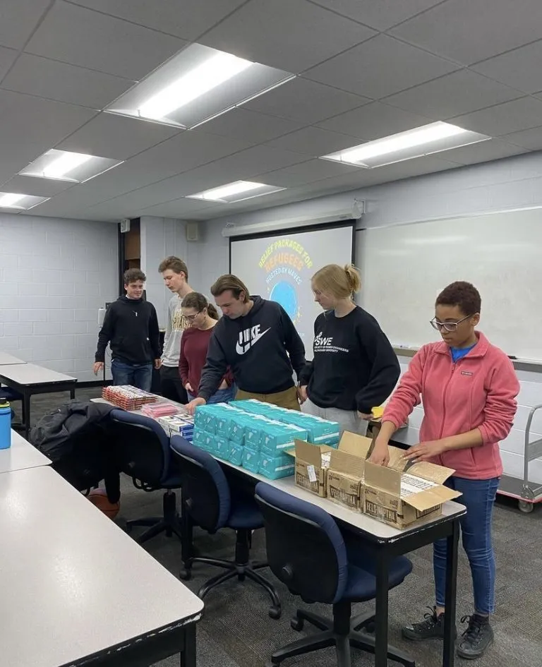 Six people stand behind a table organizing boxes and items in a classroom with a projector and whiteboard, preparing for the WAVES program activity.