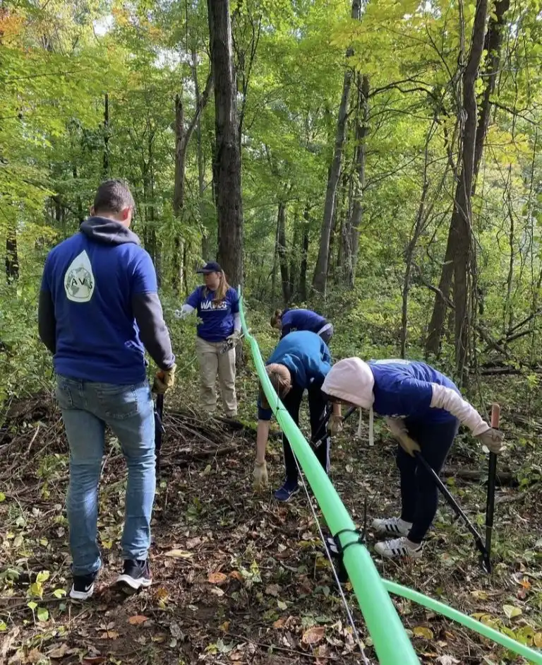 Four people in a forest install a long green WAVES pipe on the ground while wearing blue shirts and gloves.