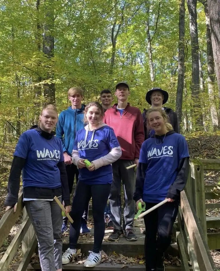 Seven people, some proudly wearing matching “WAVES” shirts, stand together on a wooden bridge in a sunlit, leafy forest.