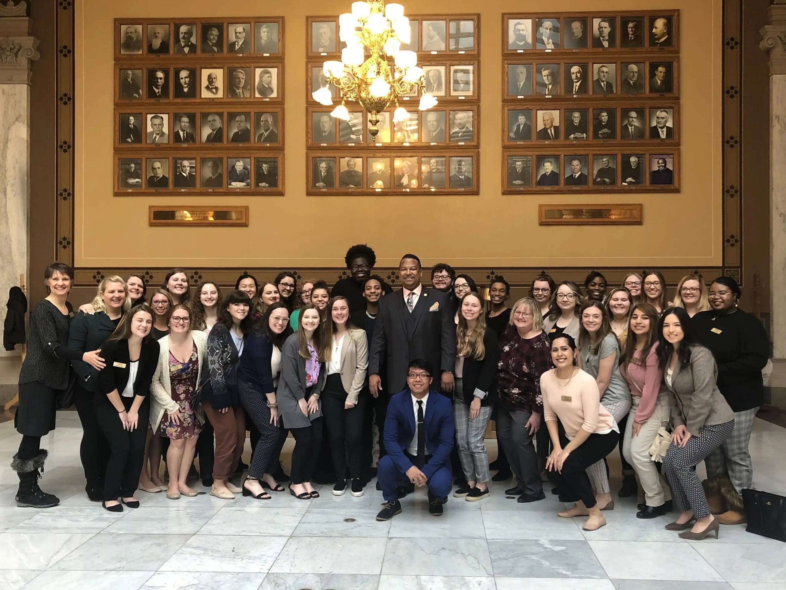 A large group of people, including Social Work students with a Minor in Direct Practice, pose and smile indoors beneath a wall of framed portraits and a chandelier.